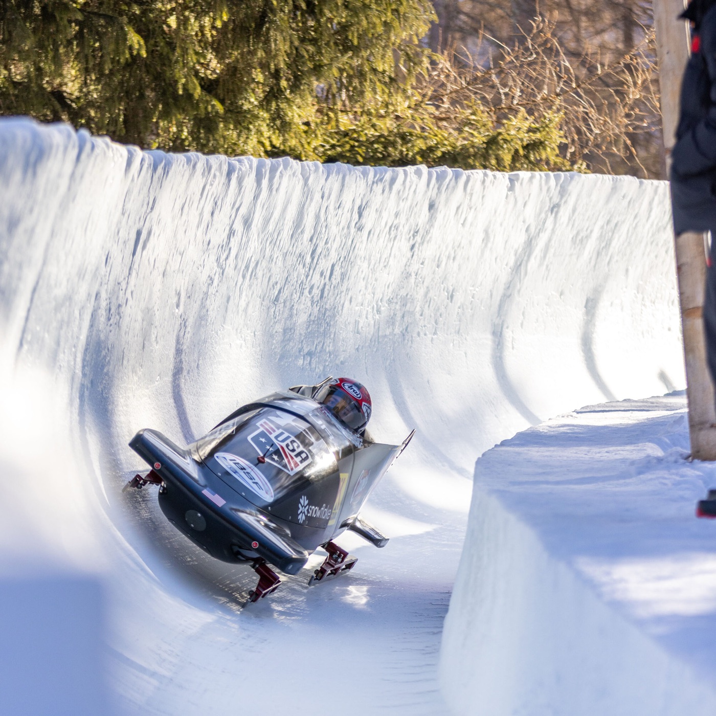 A bobsleigh leaning into a high curved wall of ice on the bob run, with fir trees in winter sunlight above