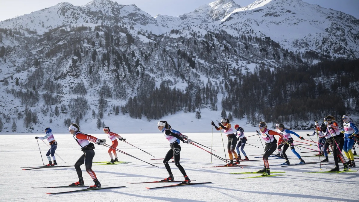 A field of cross-country skiers in race bibs skating across a frozen lake below snowy, wooded mountains