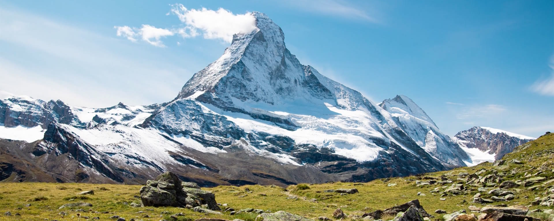 The Matterhorn under a blue summer sky seen across a grassy, rocky alpine meadow