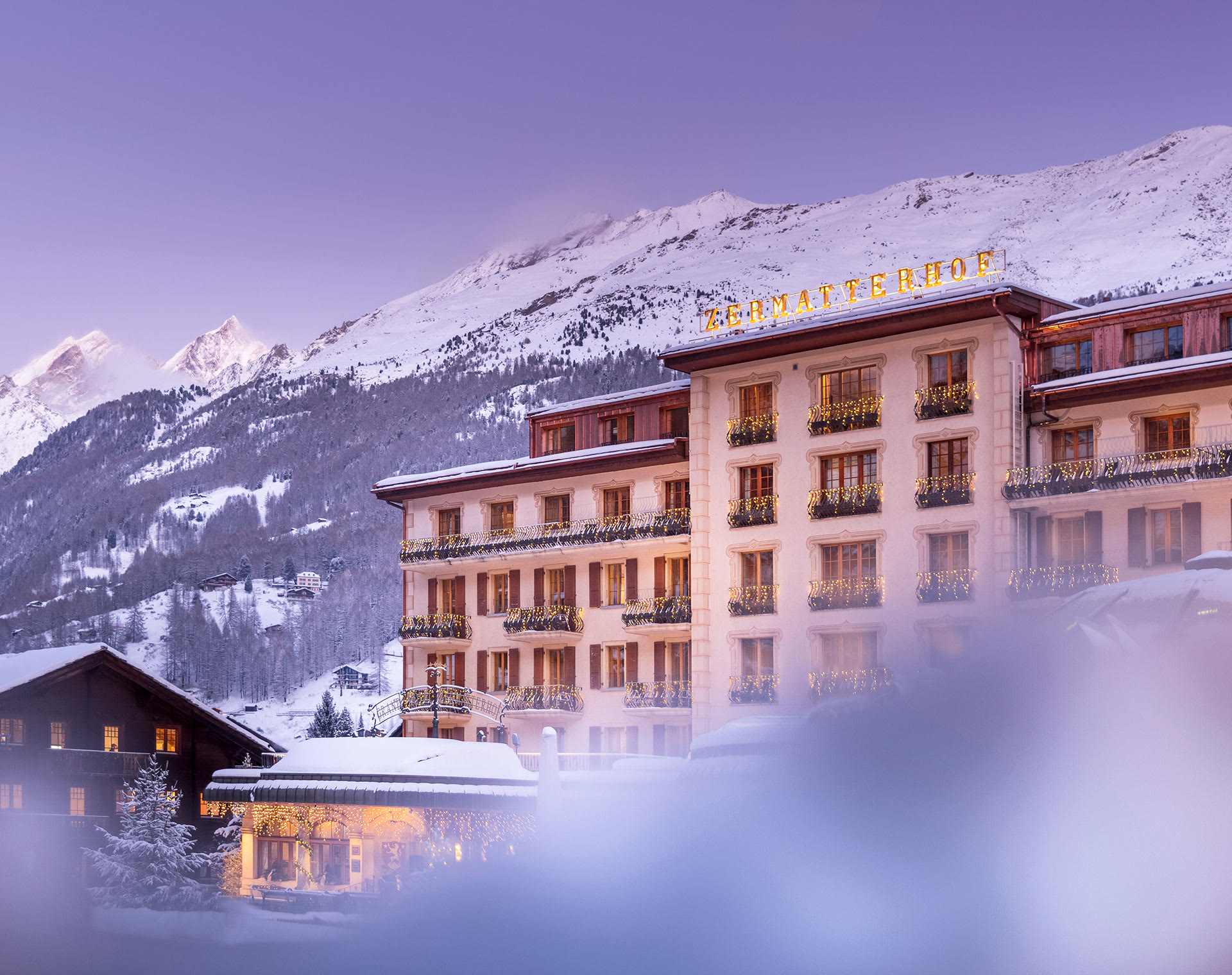 The facade of the Grand Hotel Zermatterhof at dusk in winter, with snowy mountains behind